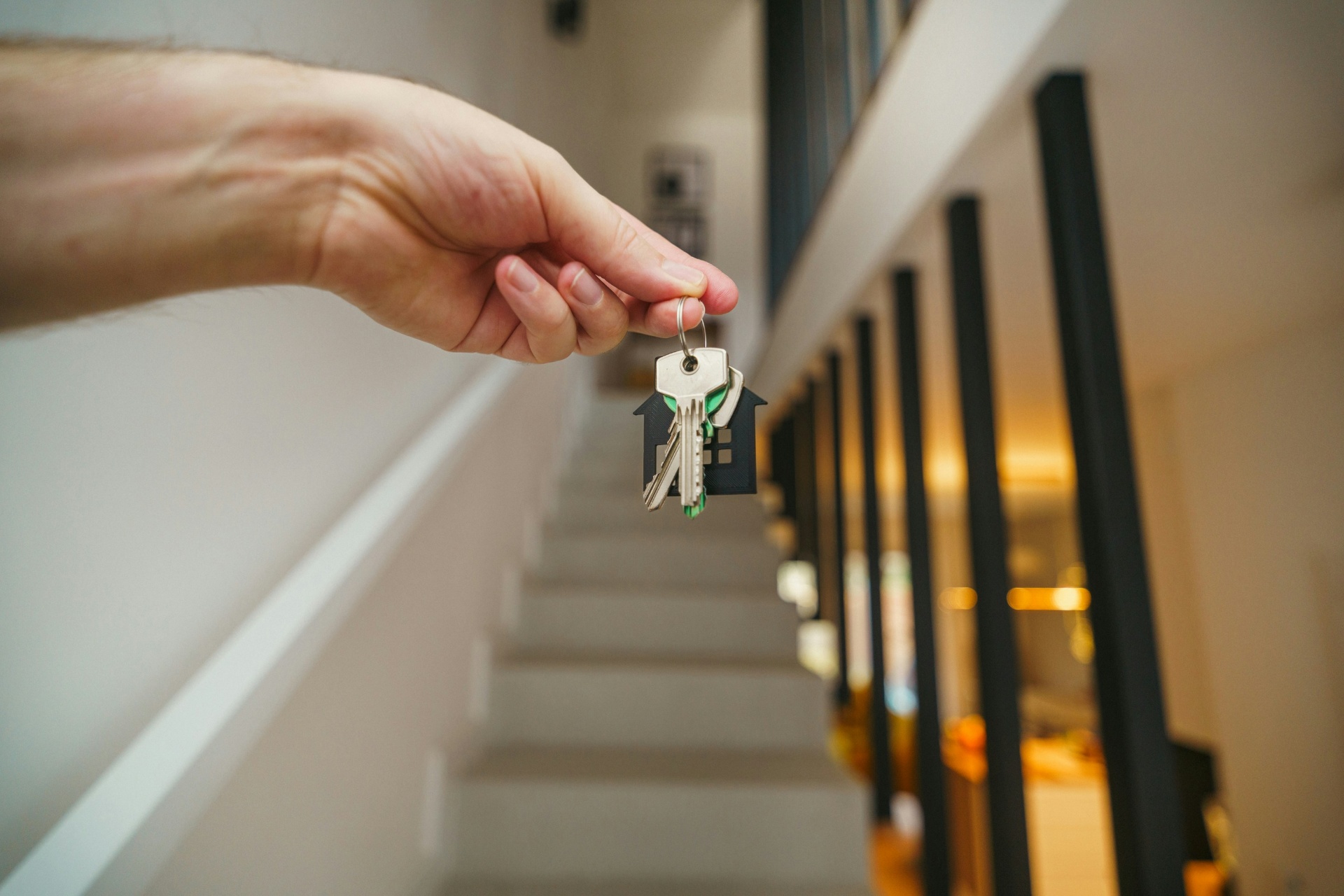 Mano sosteniendo llaves con llavero en forma de casa frente a una escalera interior